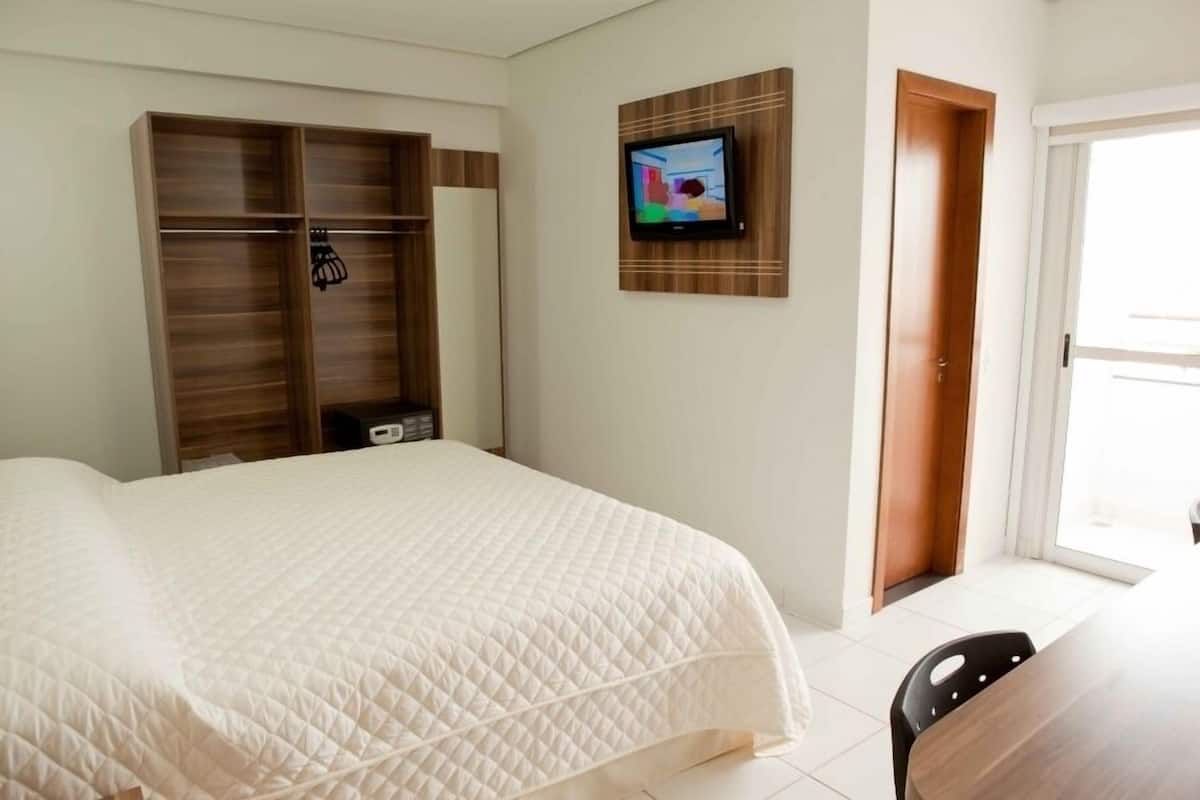 A hotel room with a large bed, a TV mounted on the wall, a wooden wardrobe, and a door leading to a balcony.