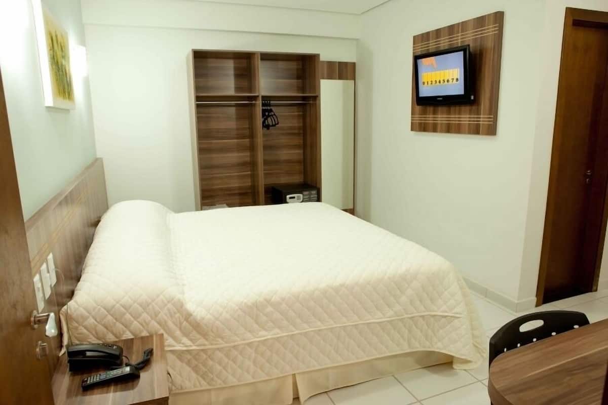 A bedroom with a bed, a wooden wardrobe, a television mounted on the wall, and a desk with a chair.