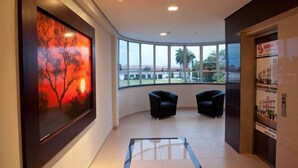 Un hall d’hôtel doté d’une grande fenêtre, de deux fauteuils noirs et d’une œuvre d’art encadrée représentant un coucher de soleil.