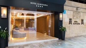 Hotel Almudainas indgang med et moderne design, der inkluderer en glasfacade, et receptionområde og en stenvæg med hotellets navn og logo.