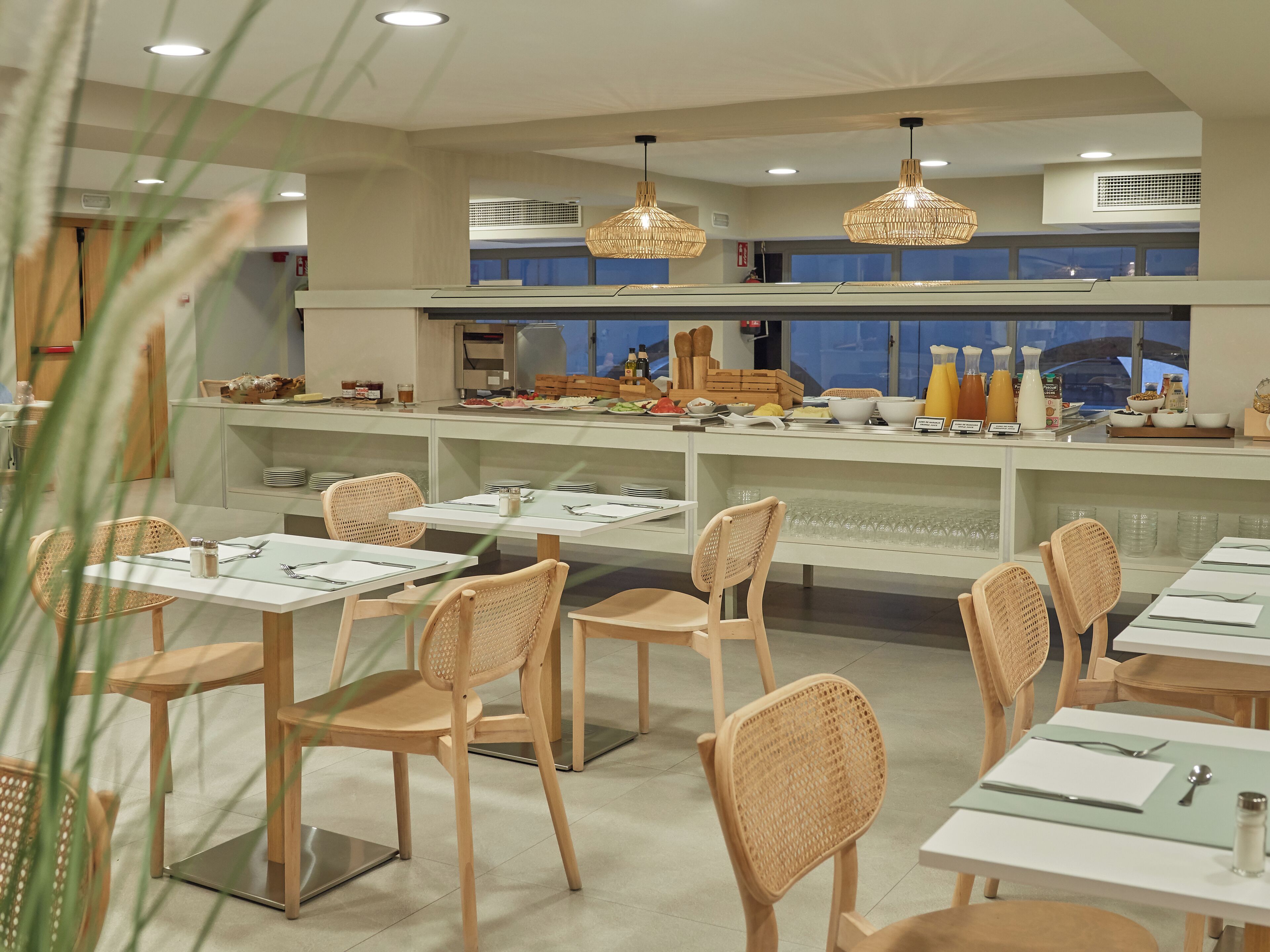 Un comedor moderno con sillas de mimbre, mesas blancas y un mostrador de buffet con diversas opciones de comida.