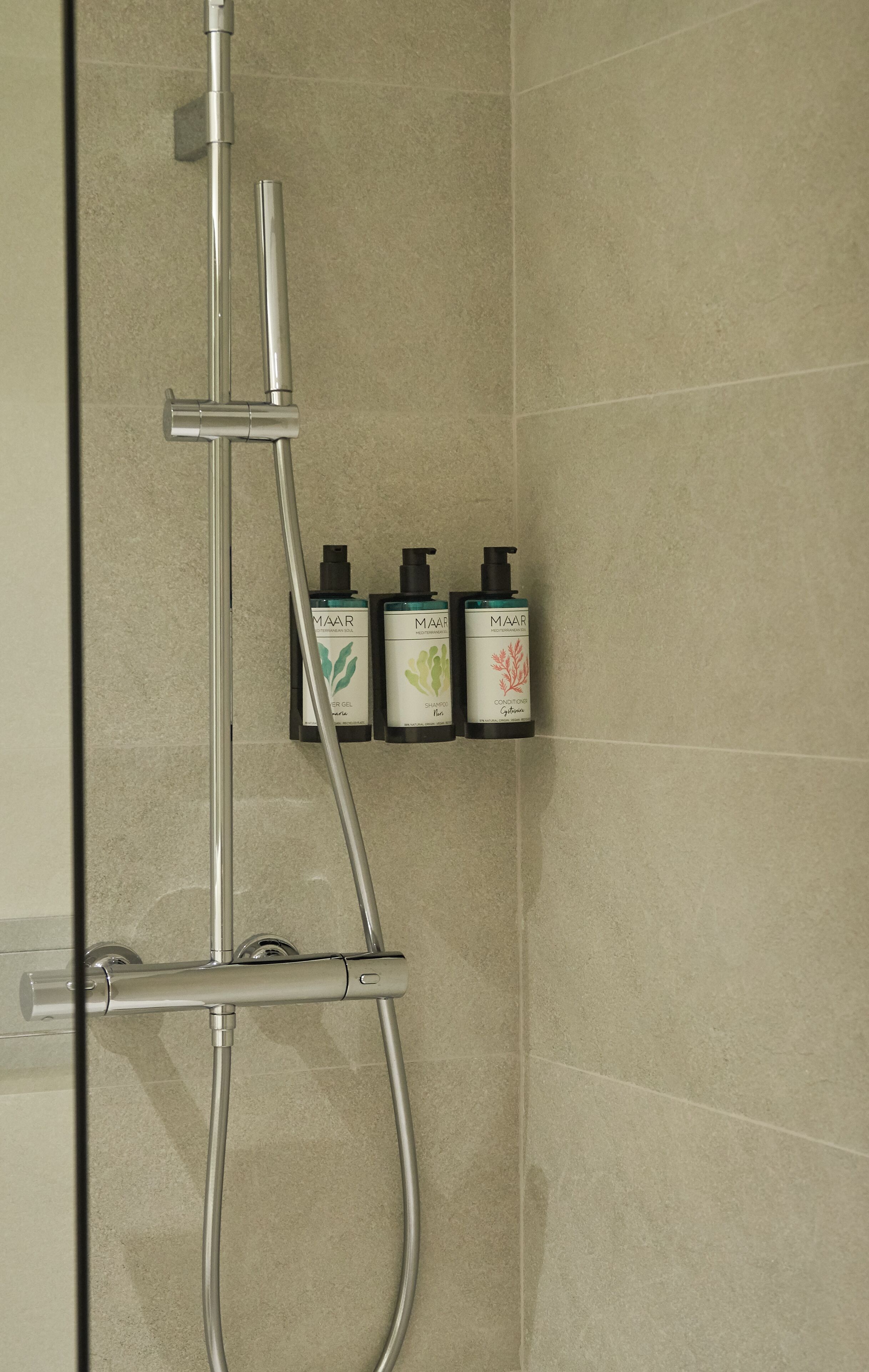 Una ducha con cabezal de ducha de mano y panel de control en la pared. Hay tres botellas de productos MAAR: champú, acondicionador y gel de ducha.