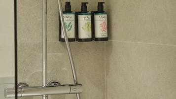 Una ducha con cabezal de ducha de mano y panel de control en la pared. Hay tres botellas de productos MAAR: champú, acondicionador y gel de ducha.