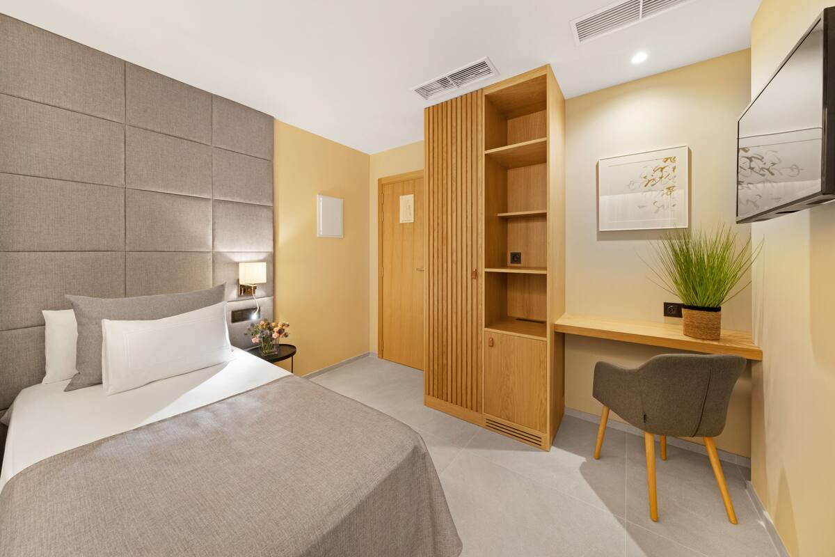 A modern hotel room with a large bed, a wooden wardrobe, a desk with a chair, and a mounted TV.