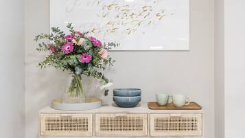 Een minimalistisch ingerichte kamer met een witte muur, een lijst met een citaat, een vaas met bloemen en een console tafel met een schaal en kopjes.