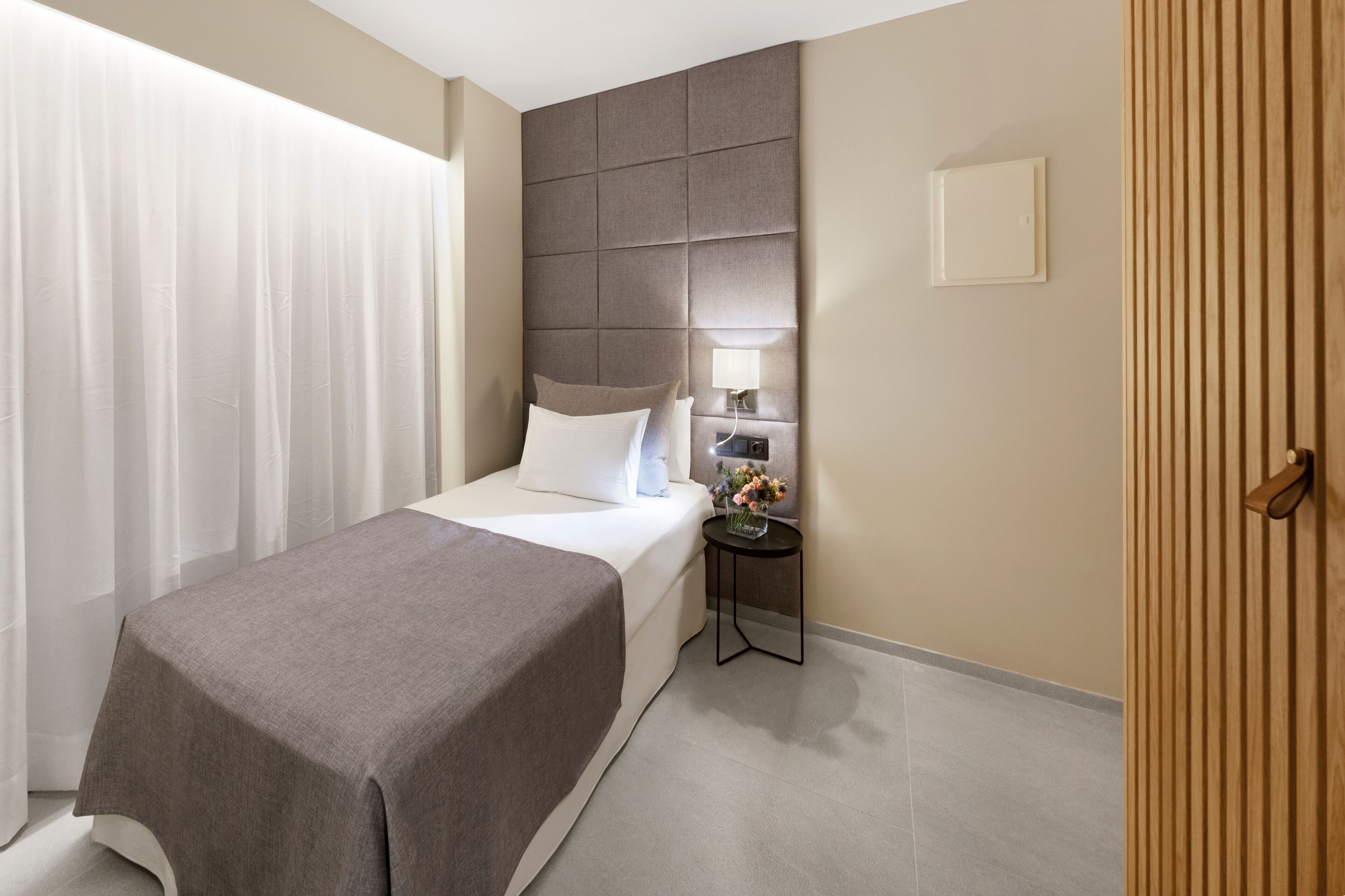 Una habitación de hotel con una cama, una mesita de noche con lámpara, una luz empotrada en la pared y una puerta de madera.