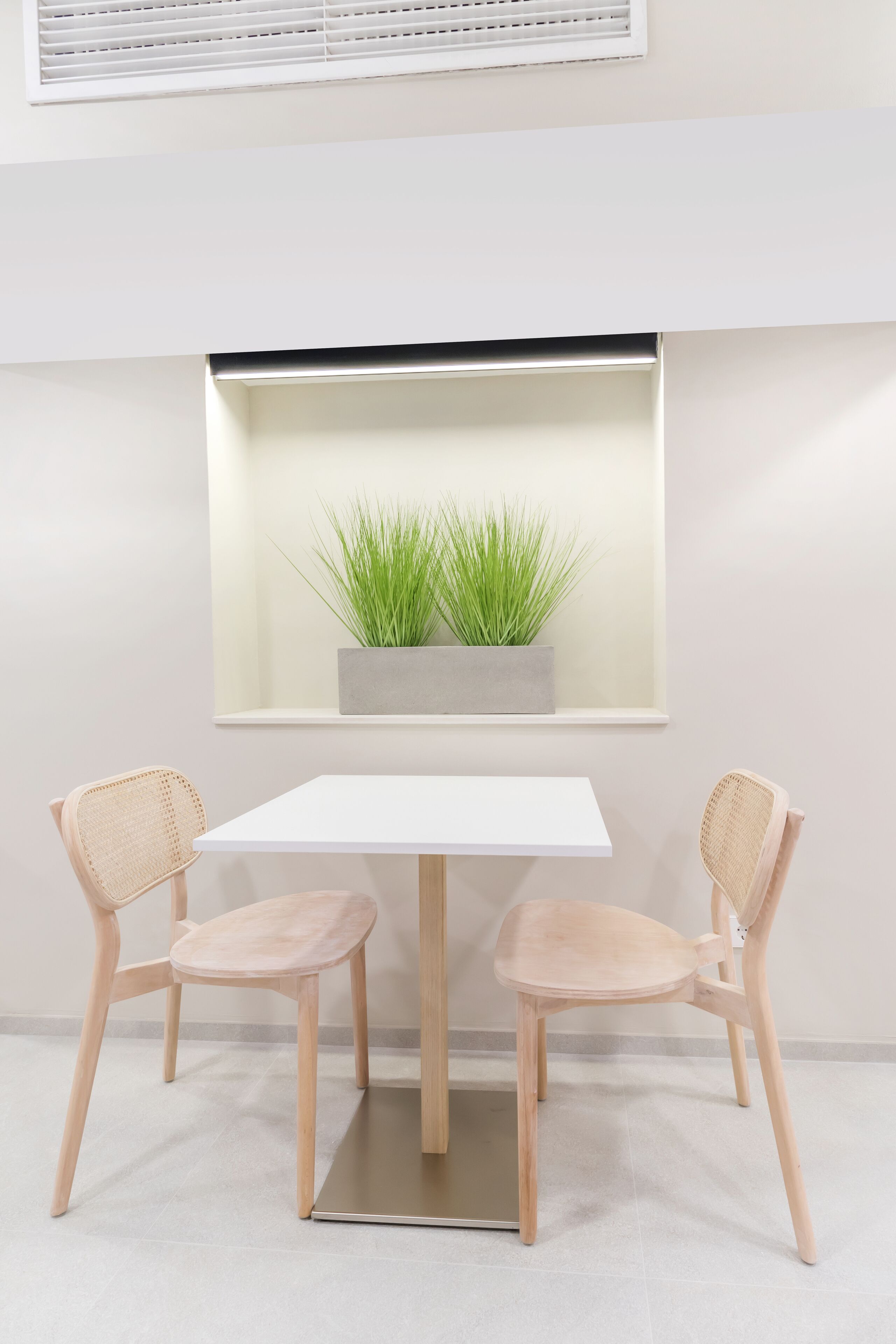 A minimalist dining area with a white table and two wicker chairs, a potted plant on a shelf above, and a window with blinds.