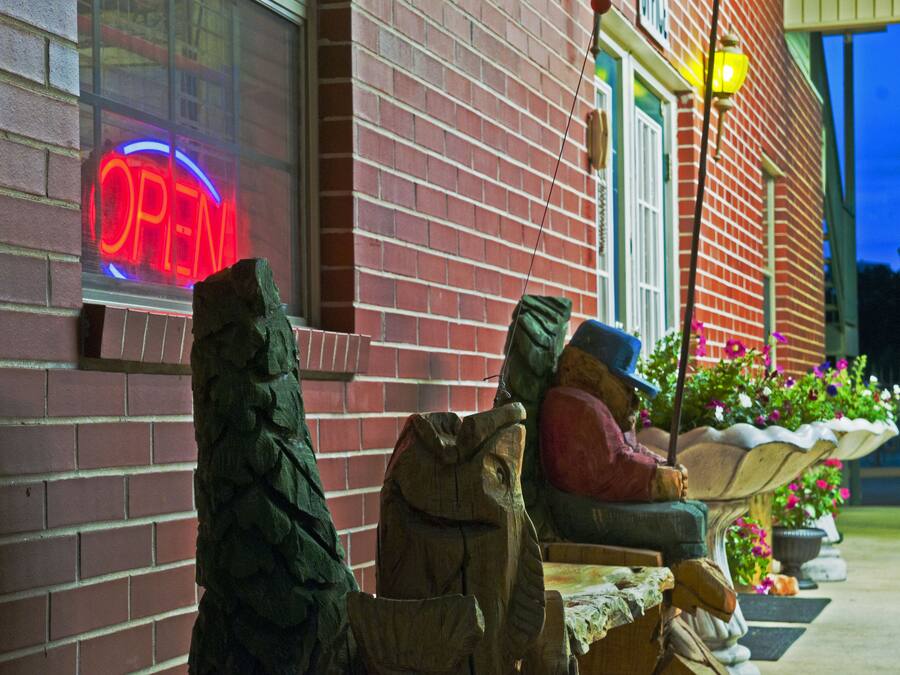 A brick building with a neon 'OPEN' sign, outdoor seating with a stone table and chairs, and a flower pot with pink flowers.