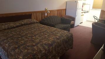 A hotel room with a large bed, a chair, a small table, a refrigerator, and a trash can.