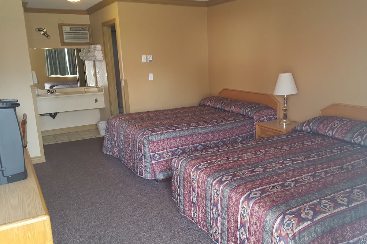 A hotel room with two beds, a small desk, a lamp, a mirror, and a sink.