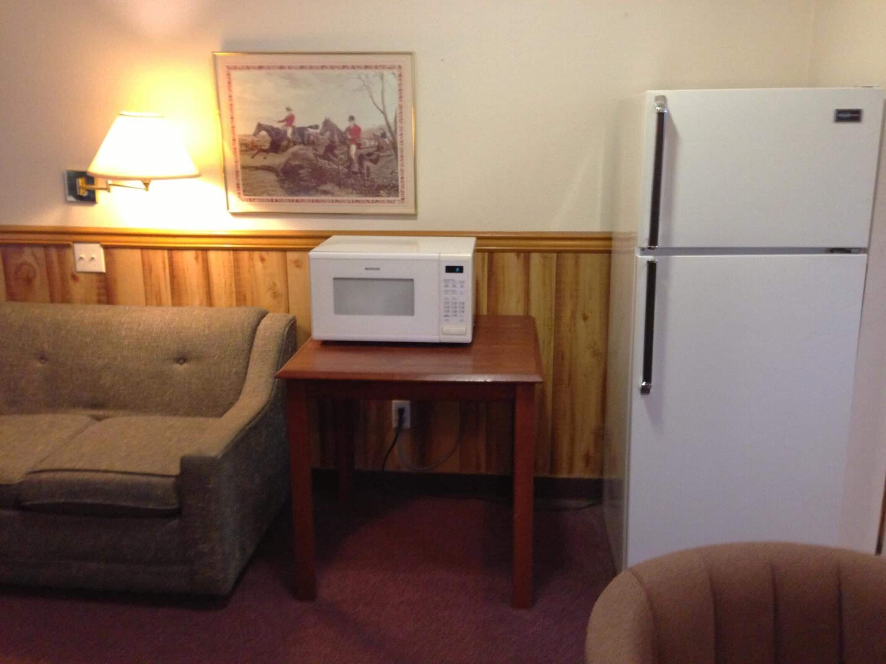 A room with a sofa, a table with a microwave, a refrigerator, and a wall-mounted lamp.