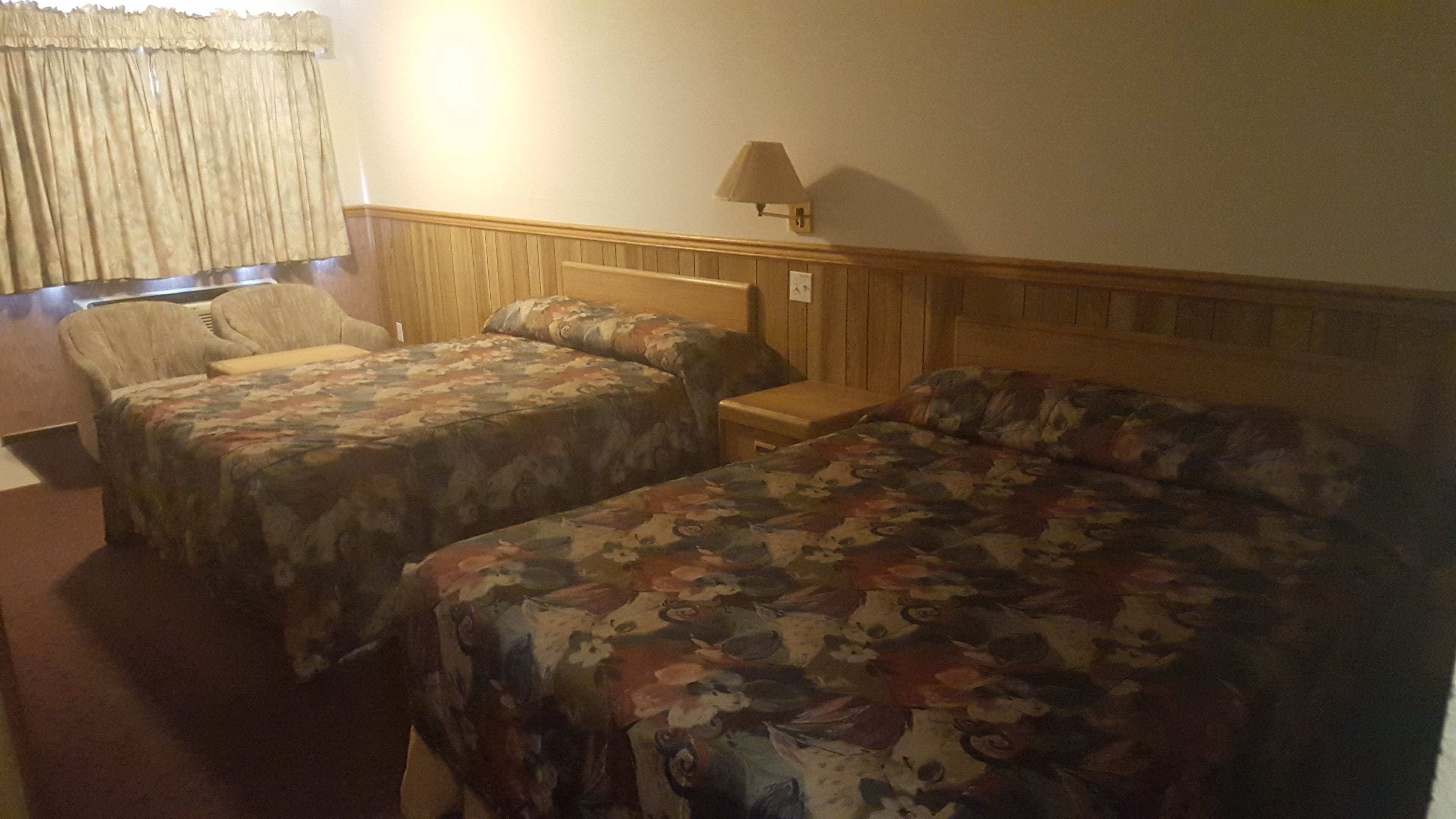 A hotel room with two beds, a nightstand, a lamp, and a chair.