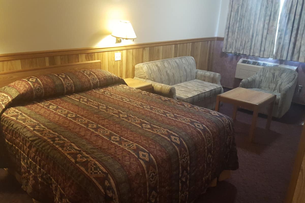 A hotel room with a large bed, two armchairs, a small table, a lamp, and an air conditioning unit.