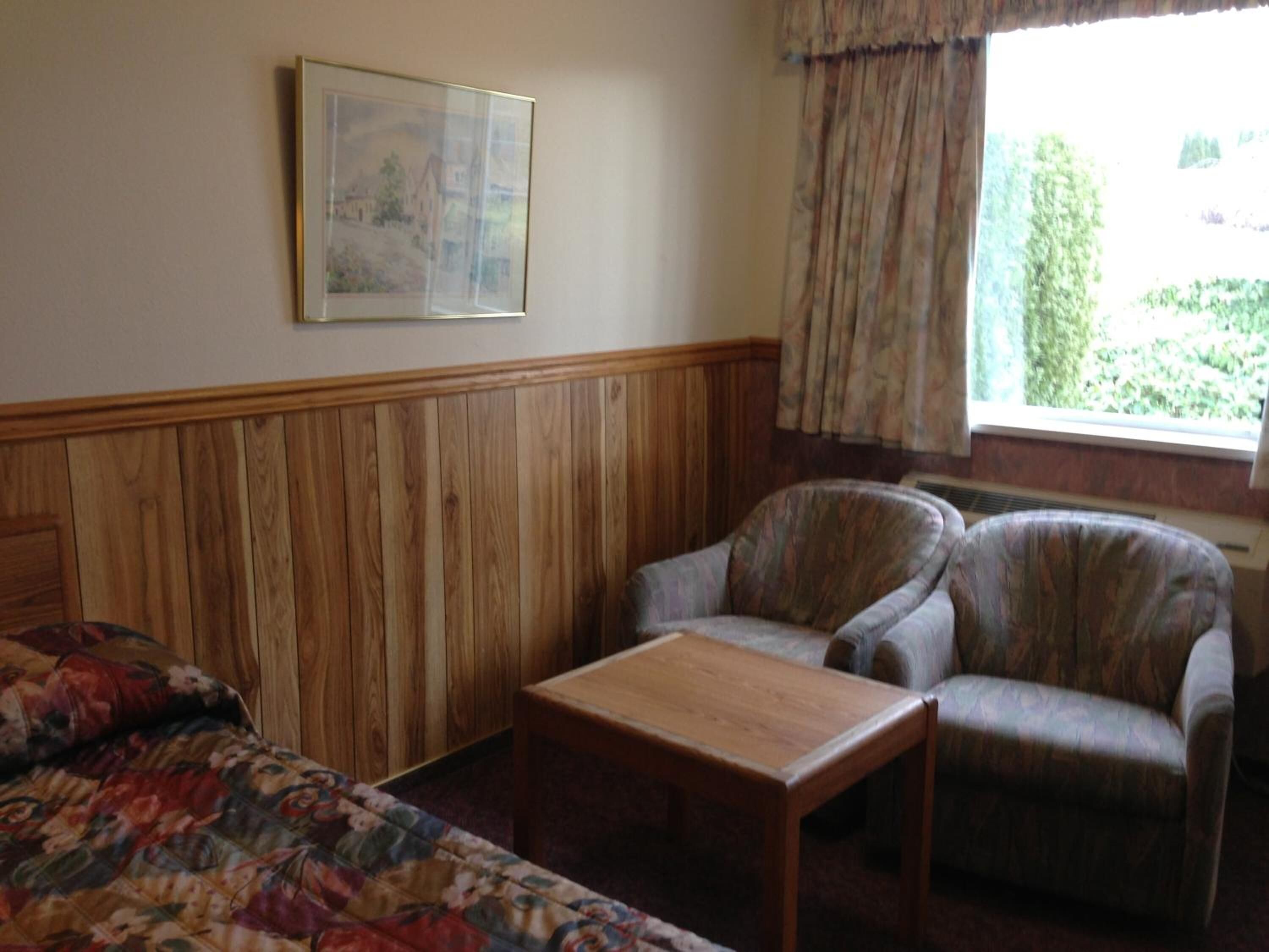 A room with a wooden paneled wall, a bed with a floral bedspread, two armchairs, a small wooden table, and a window with curtains.