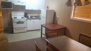 Una cocina compacta con electrodomésticos blancos, una mesa de madera y una ventana con persianas.