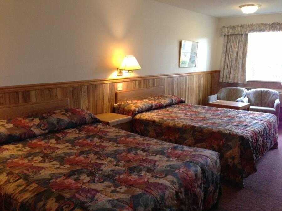 Una habitación de hotel con dos camas, una mesita de noche, una silla y un ventanal con cortinas.