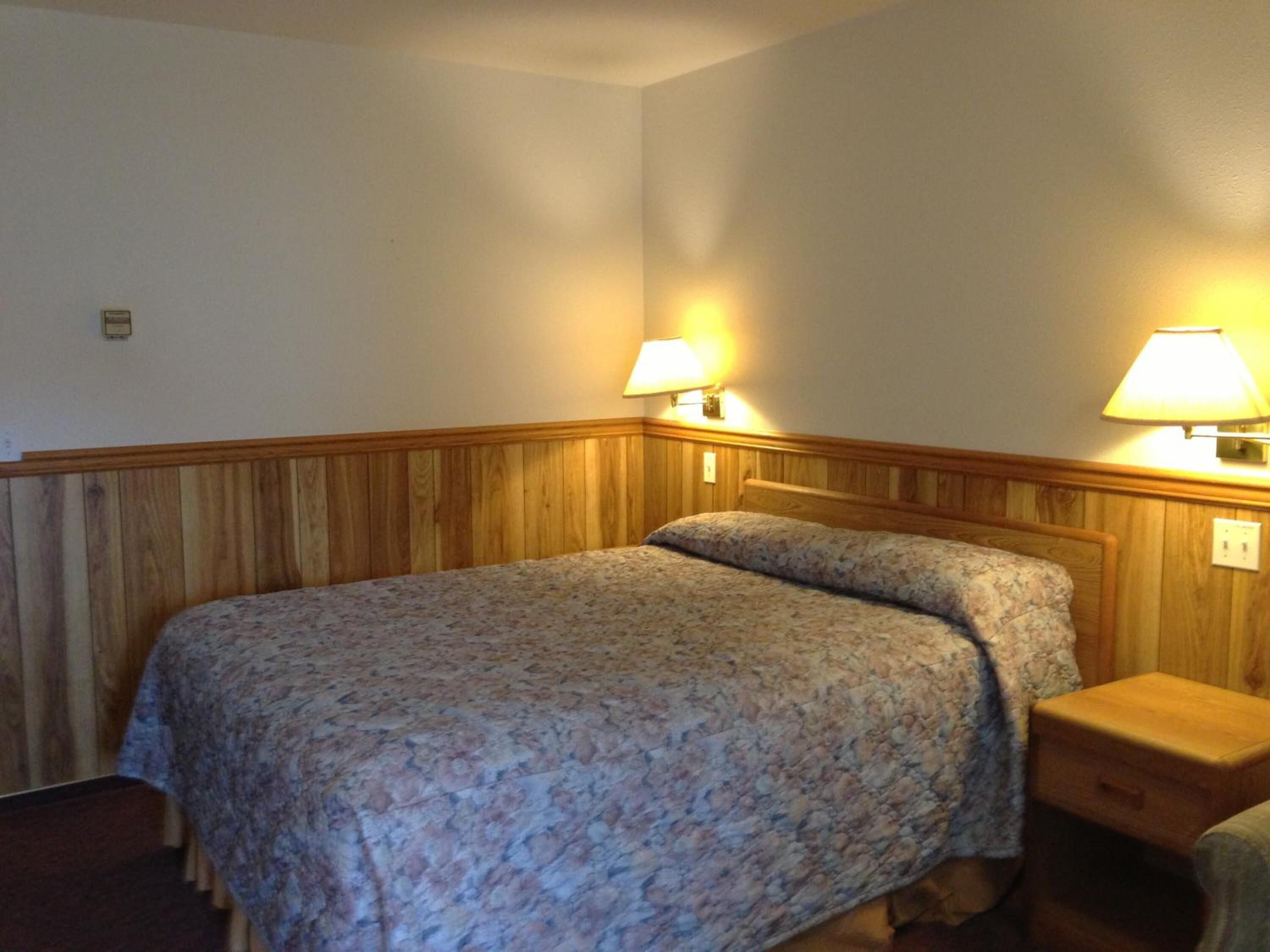 A single bed with a floral bedspread in a room with wooden paneling on the walls and a nightstand with a lamp.