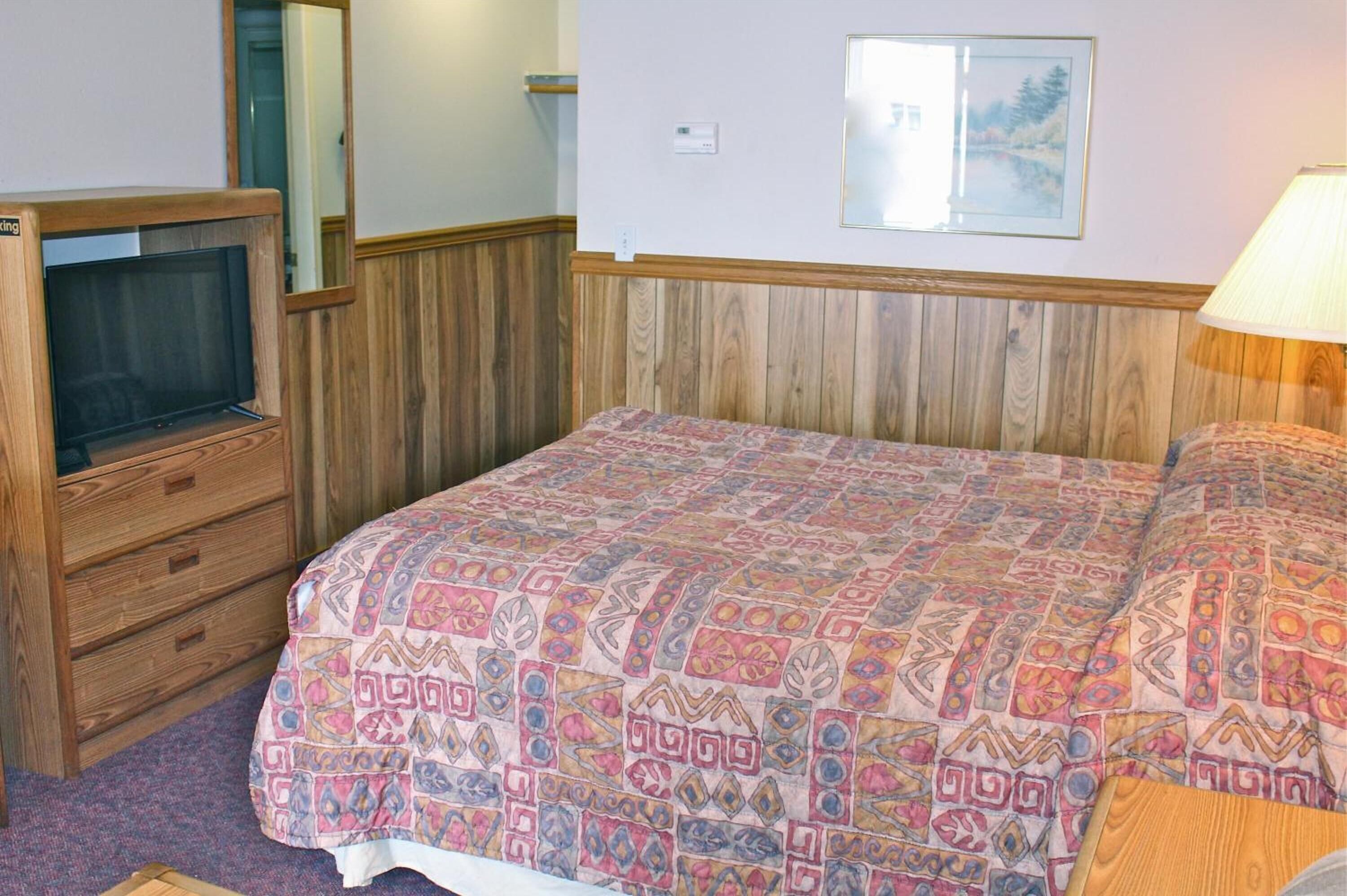 A bedroom with a wooden headboard, a bed with a patterned bedspread, a dresser with a mirror, and a wall-mounted thermostat.