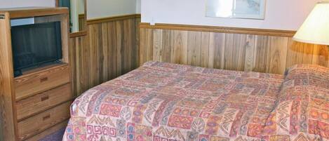 A bedroom with a wooden headboard, a bed with a patterned bedspread, a dresser with a mirror, and a wall-mounted thermostat.