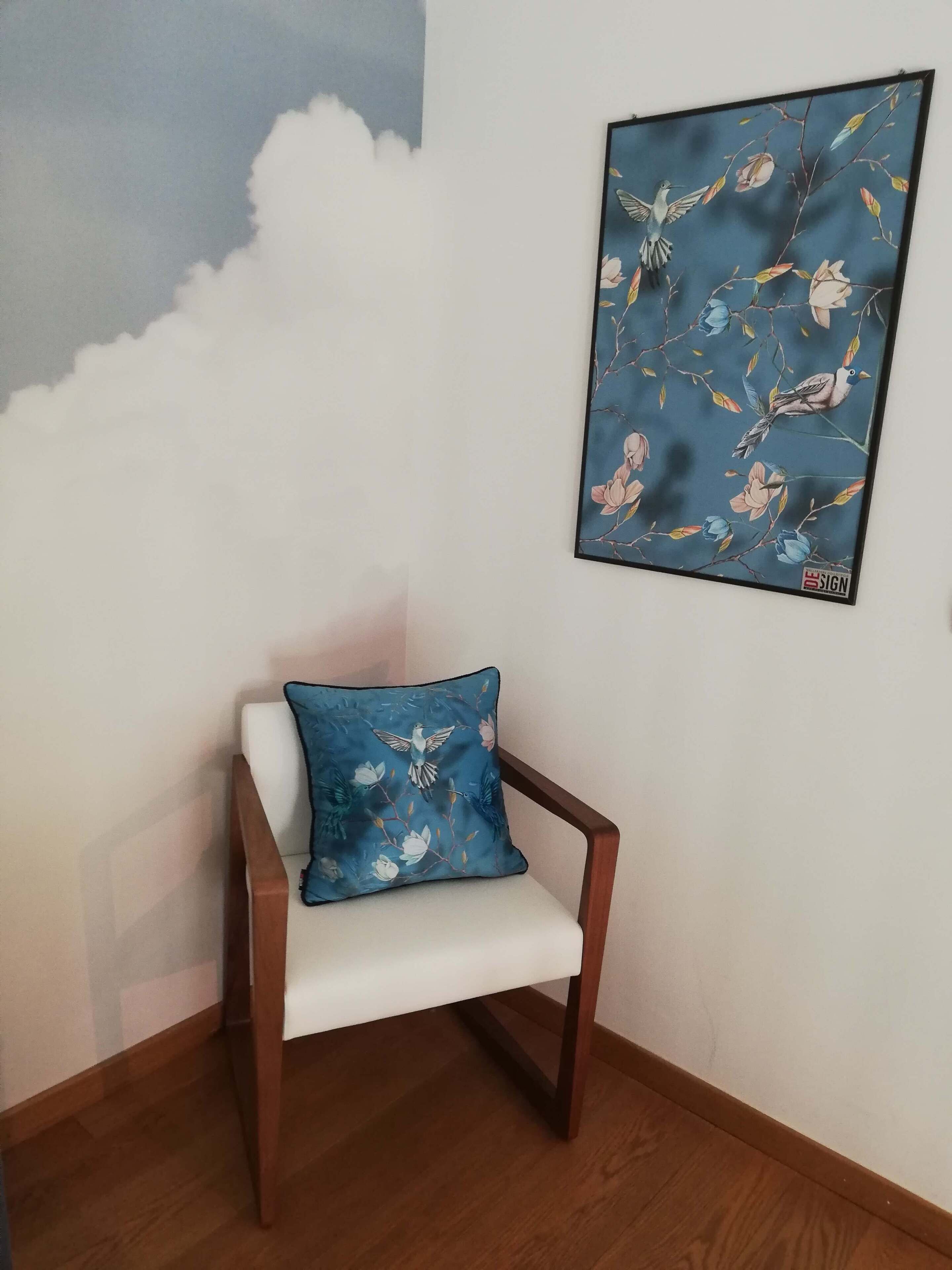 A chair with a floral cushion against a wall with a framed artwork depicting birds and flowers.