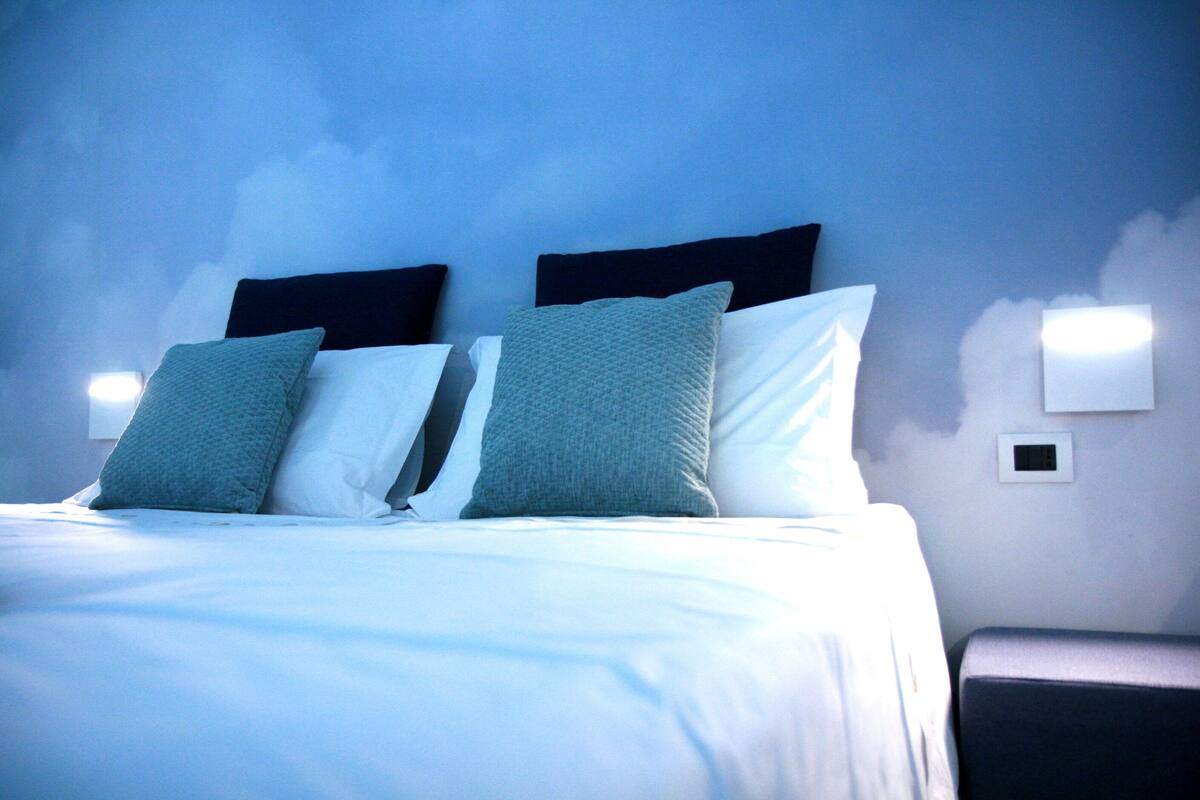 A neatly made bed with white and teal pillows, a dark headboard, and a wall-mounted light fixture.
