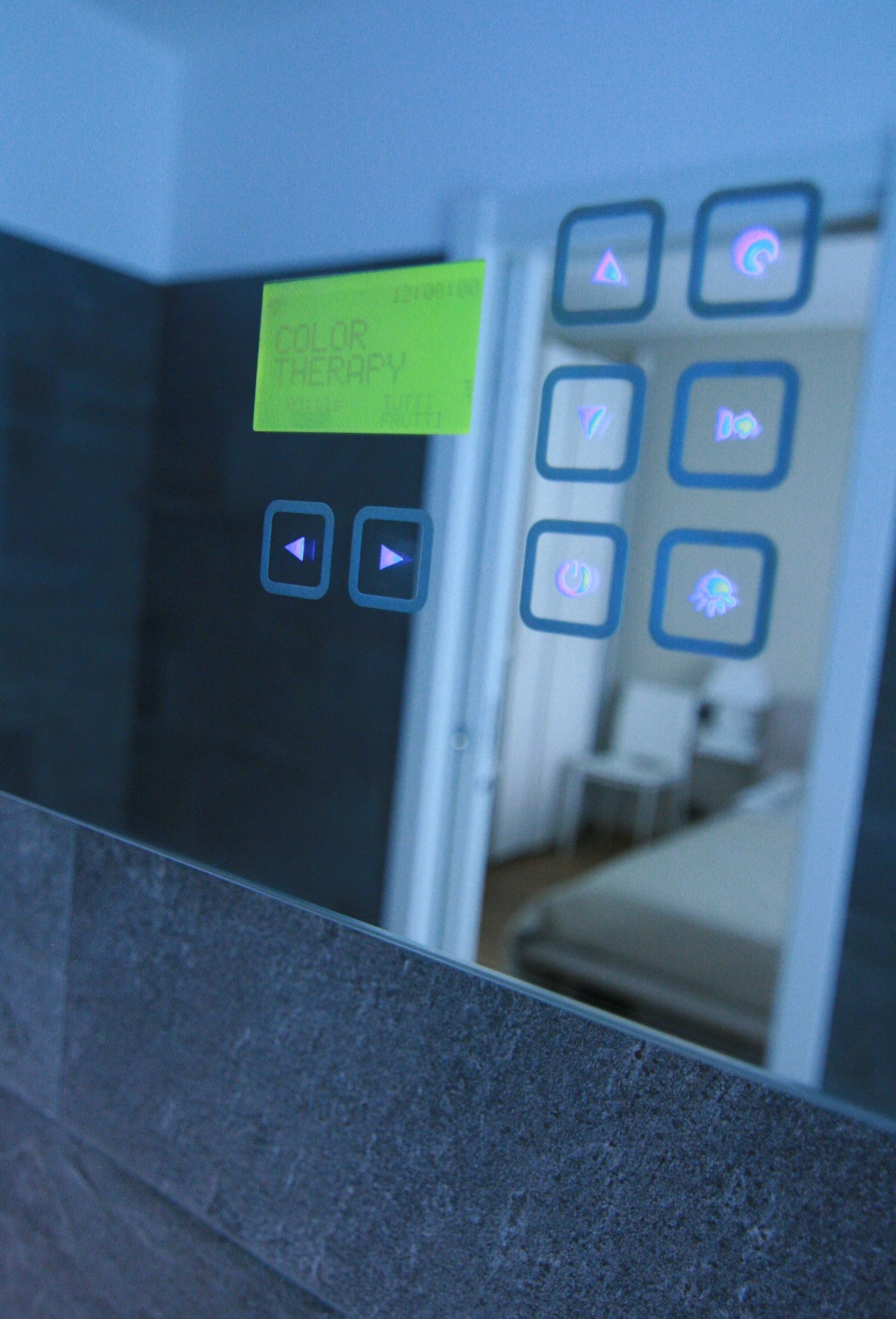 Un bagno con uno specchio che riflette una stanza. Un post-it sullo specchio recita "COLOR THERAPY". Sono presenti dei pulsanti sulla parete, tra cui un pulsante di accensione e un pulsante con un simbolo a forma di fiore.