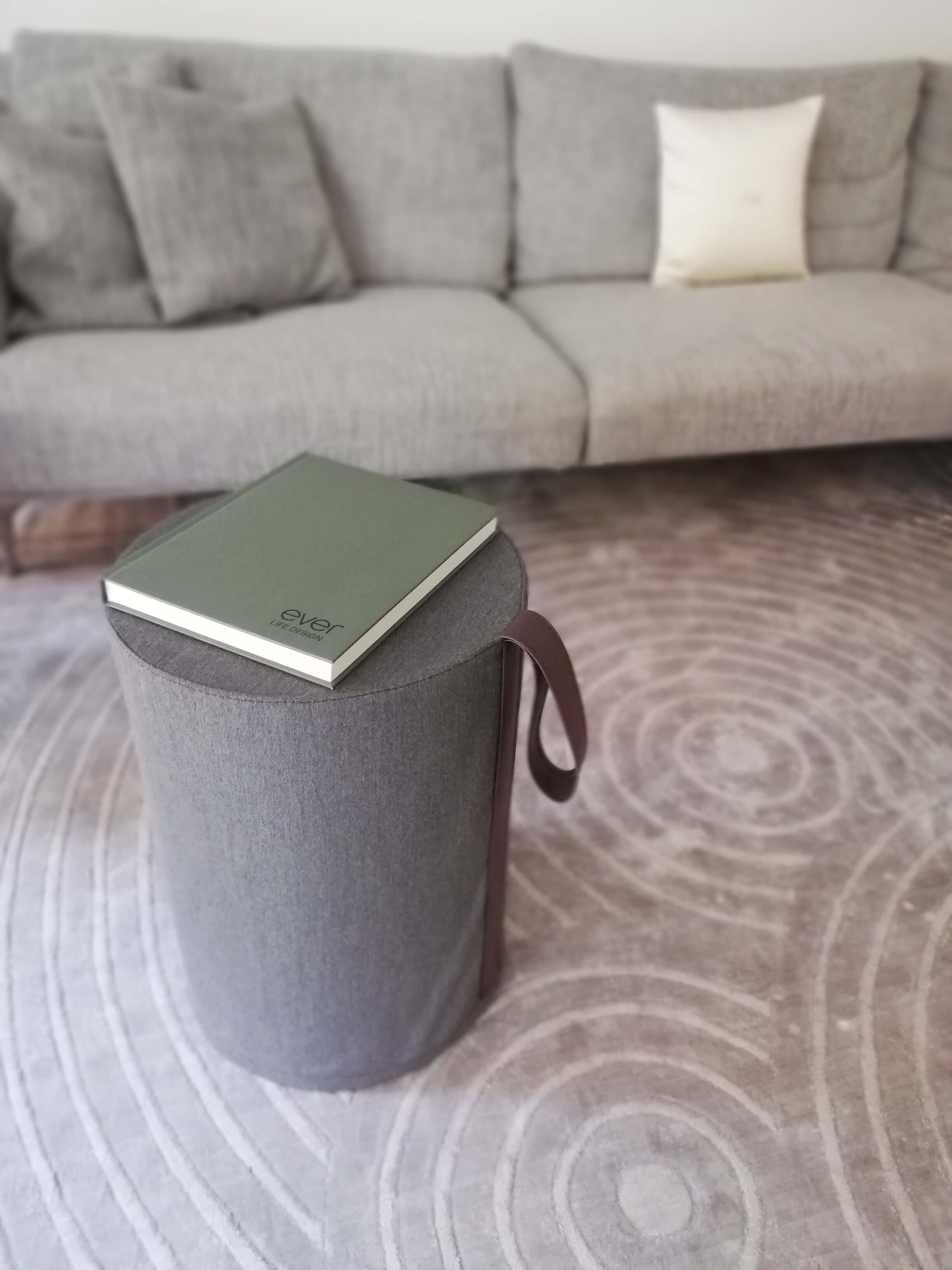 Un poggiapiedi cilindrico grigio con un libro sopra e un manico marrone, posto su un tappeto decorato.