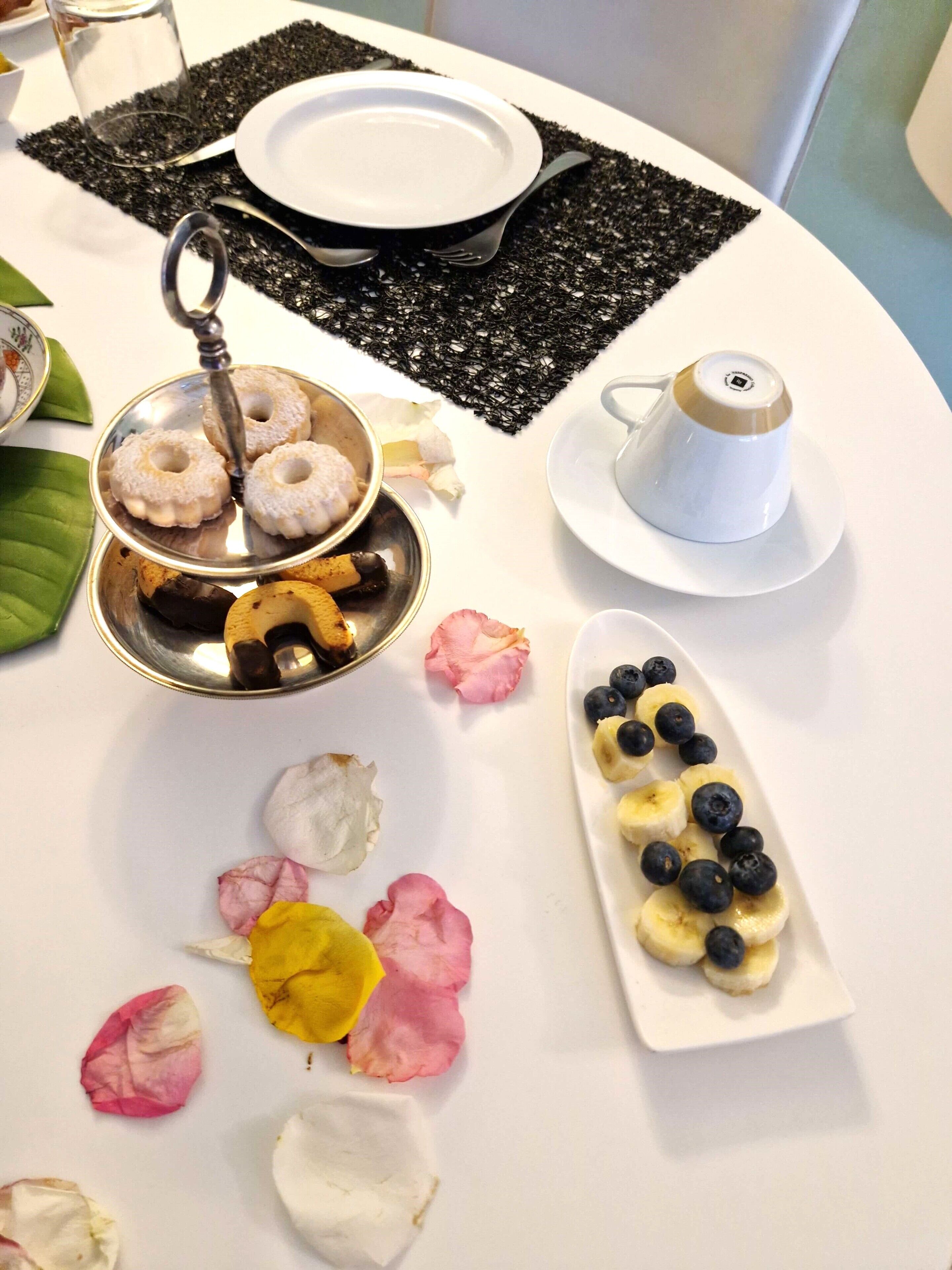 Une table avec une assiette blanche, un bol en métal contenant des beignets, une tasse blanche et une assiette avec des bananes et des myrtilles.
