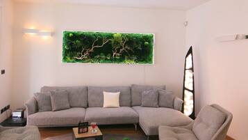 A modern living room with a grey sectional sofa, a round coffee table, and a grey armchair. There is a green wall art piece above the sofa and a geometric light fixture on the ceiling.