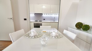 A modern kitchen with white cabinets, a stainless steel stove, and a dining area with a white table, chairs, and a glass sugar bowl. - Experience Design Bed & Show (Milan)