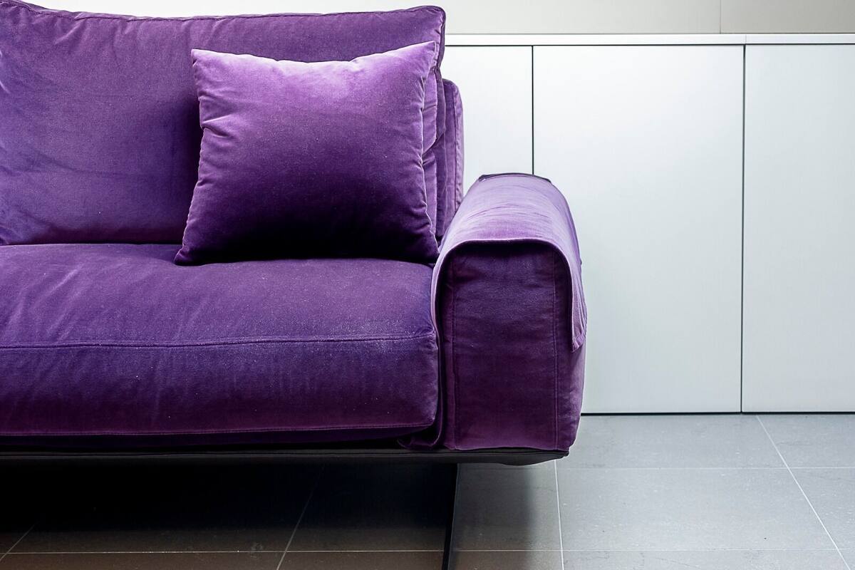 A purple sofa with a matching cushion in a modern room with white cabinets and dark tiled flooring.