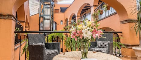 A round table with wicker chairs and a vase of flowers, set in a courtyard with arches and potted plants.