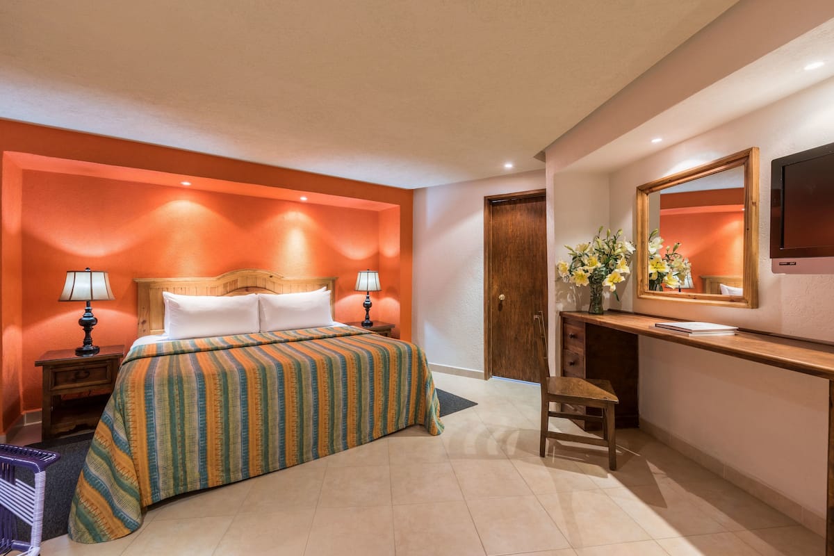 Una habitación de hotel con cama, mesitas de noche, un escritorio con silla, un espejo y un televisor.
