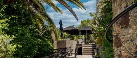 An outdoor patio with tables and chairs, surrounded by lush greenery and a stone wall.