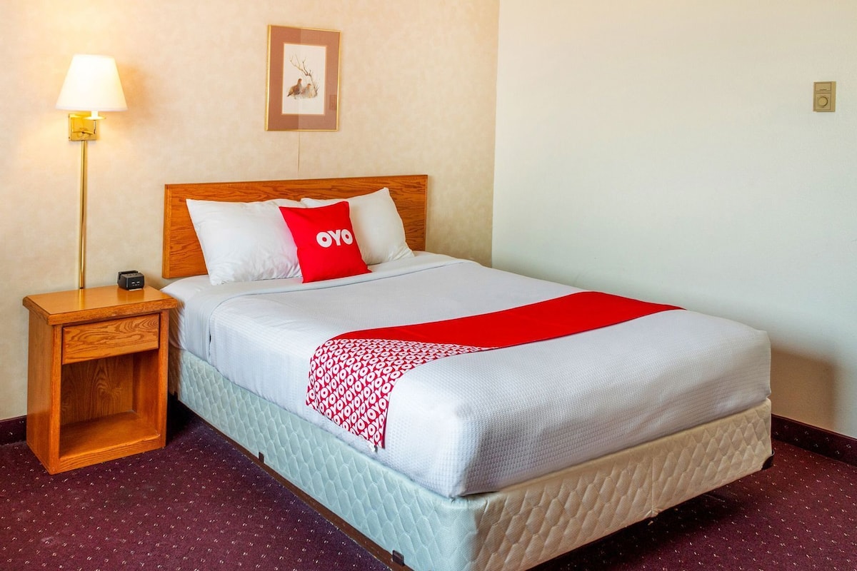 A neatly made bed with a red and white blanket, a wooden headboard, a nightstand with a lamp, and a framed picture on the wall.