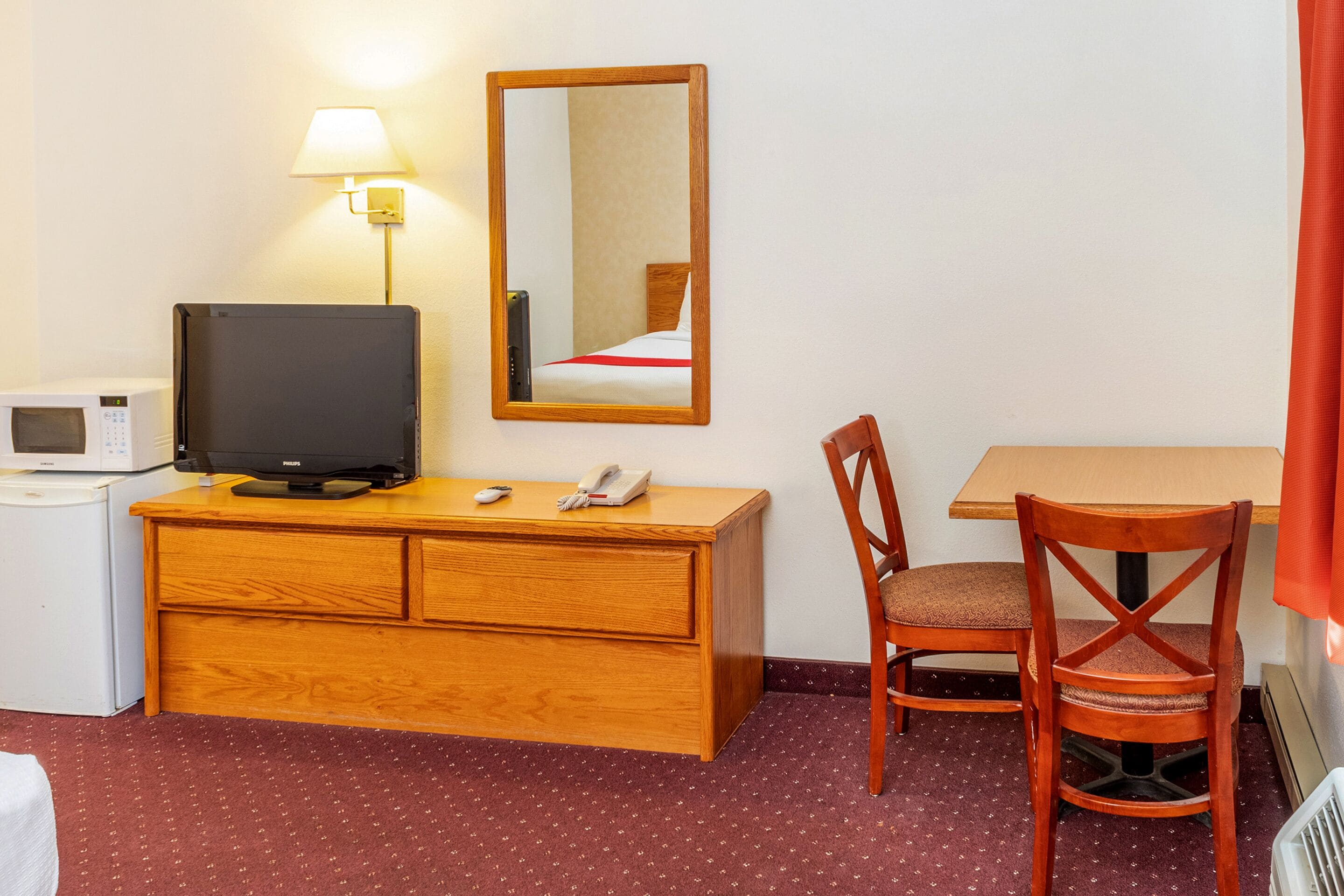 A hotel room with a wooden desk, a television, a telephone, a lamp, a mirror, a microwave, and a red carpeted floor.