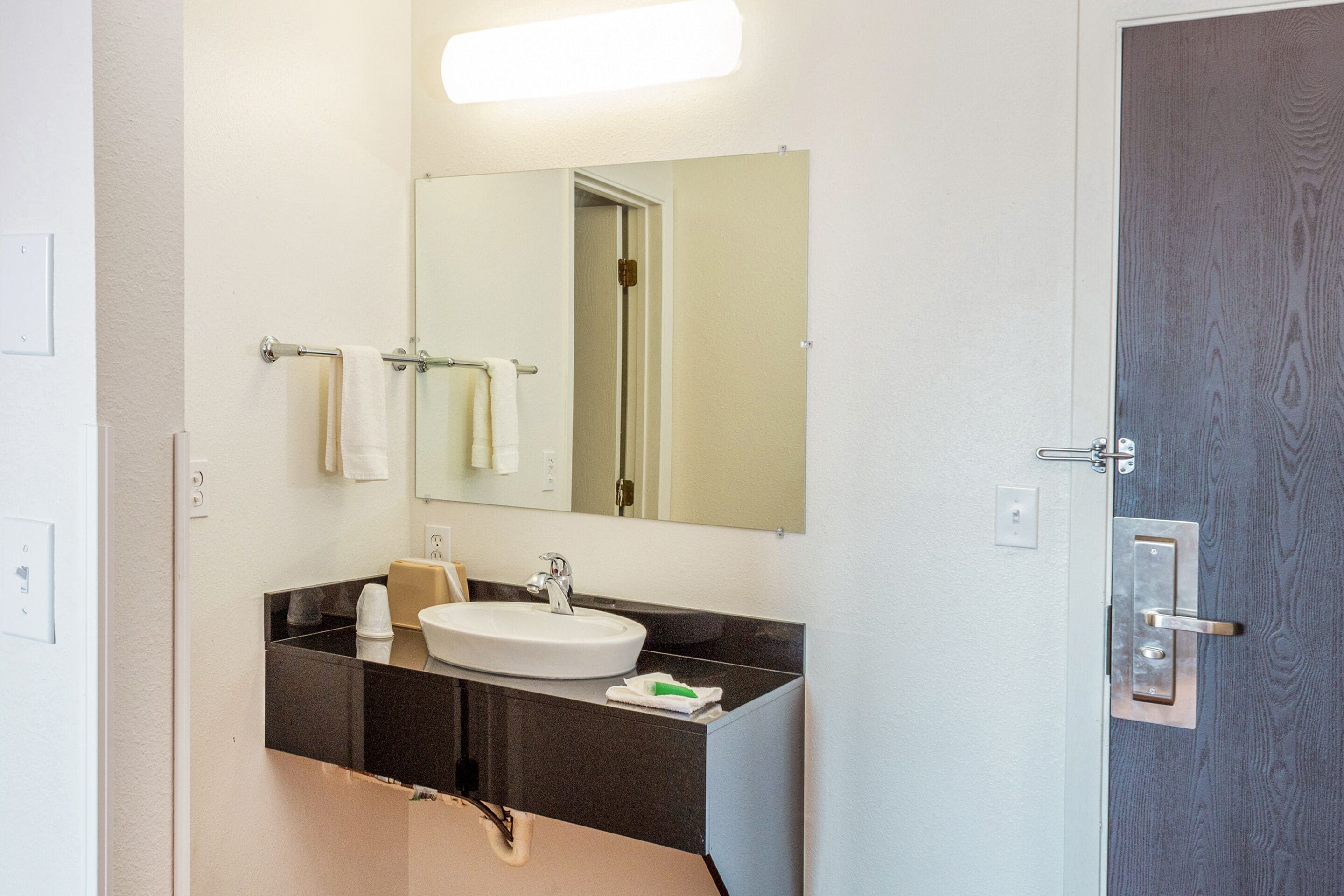 A bathroom with a white sink, dark countertop, and a mirror above it. There are towels hanging on a rack and a door with a handle.