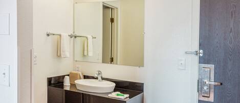 A bathroom with a white sink, dark countertop, and a mirror above it. There are towels hanging on a rack and a door with a handle.