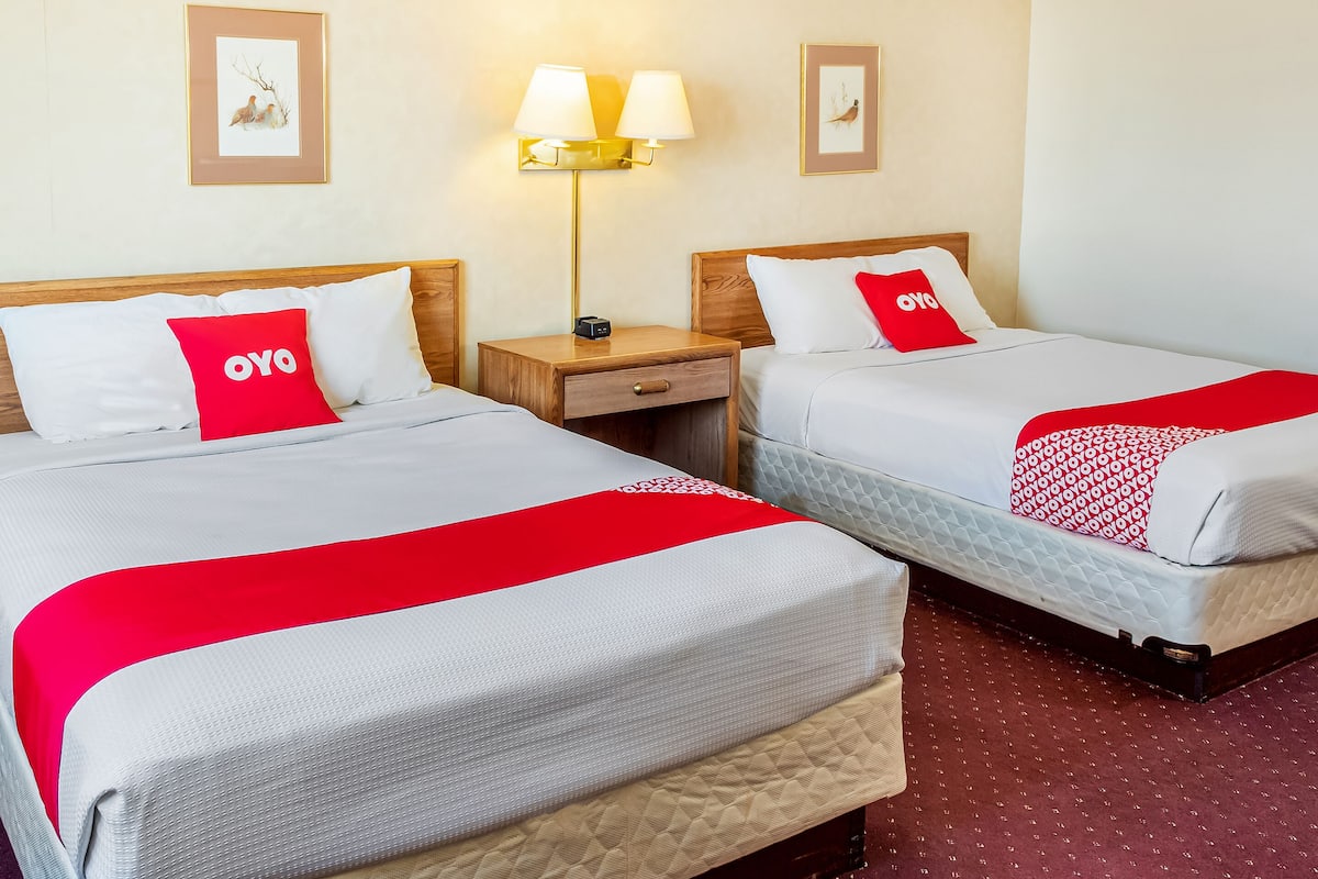 Two double beds with white bedding and red accents, a bedside table with a lamp, and framed artwork on the walls.