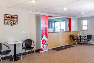 Zona de recepción de un hotel con un cartel rojo, un tablón de anuncios y dos mesas con sillas.