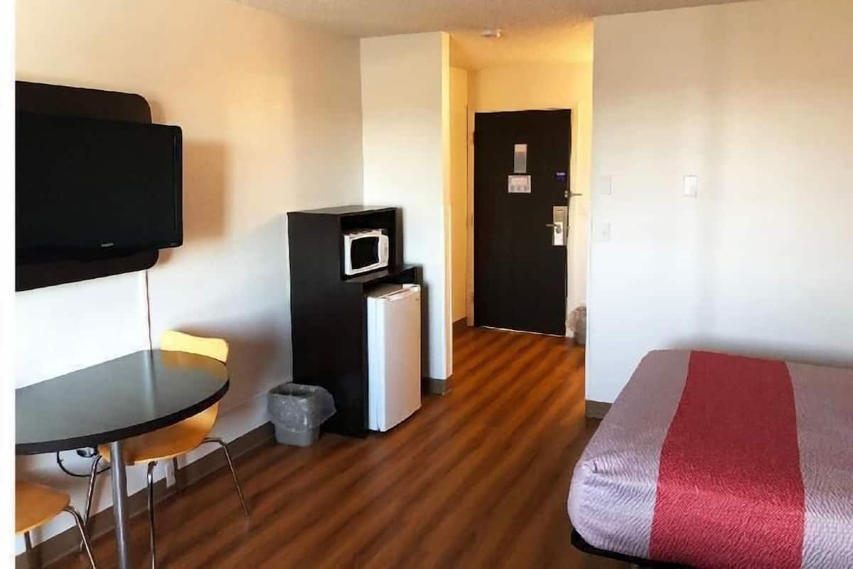 A hotel room with a bed, a desk with a chair, a television, a microwave, and a trash bin.