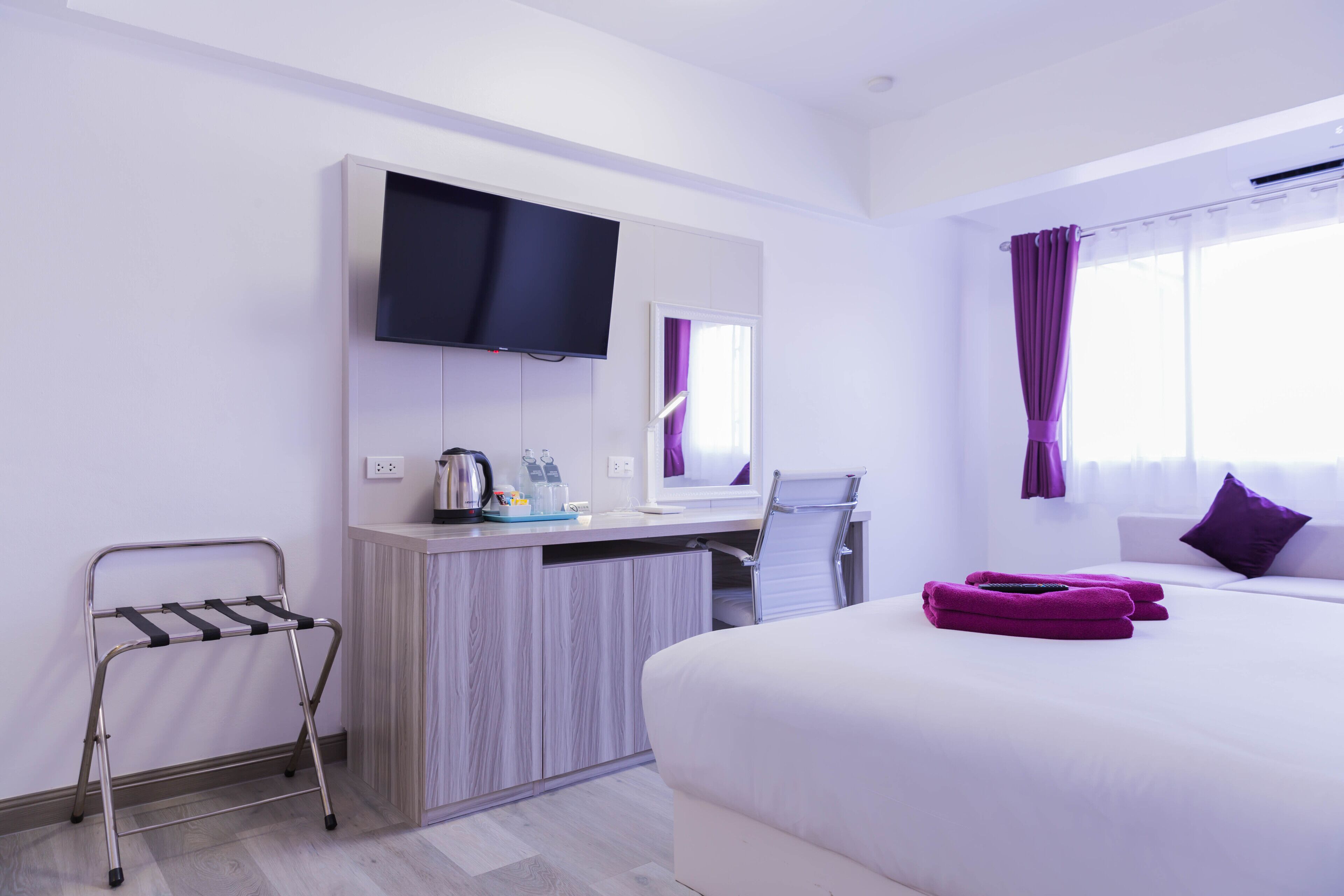 A hotel room with a bed, a desk with a flat-screen TV, a chair, a small table with a kettle, and a window with purple curtains.