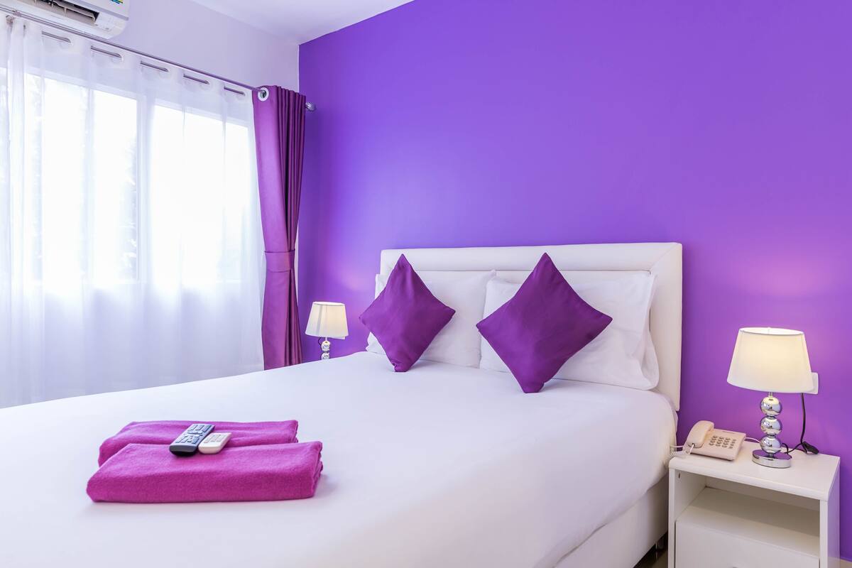 Habitación de hotel con cama, almohadas moradas, cabecera blanca, mesita de noche con lámpara y teléfono, y ventana con cortinas.