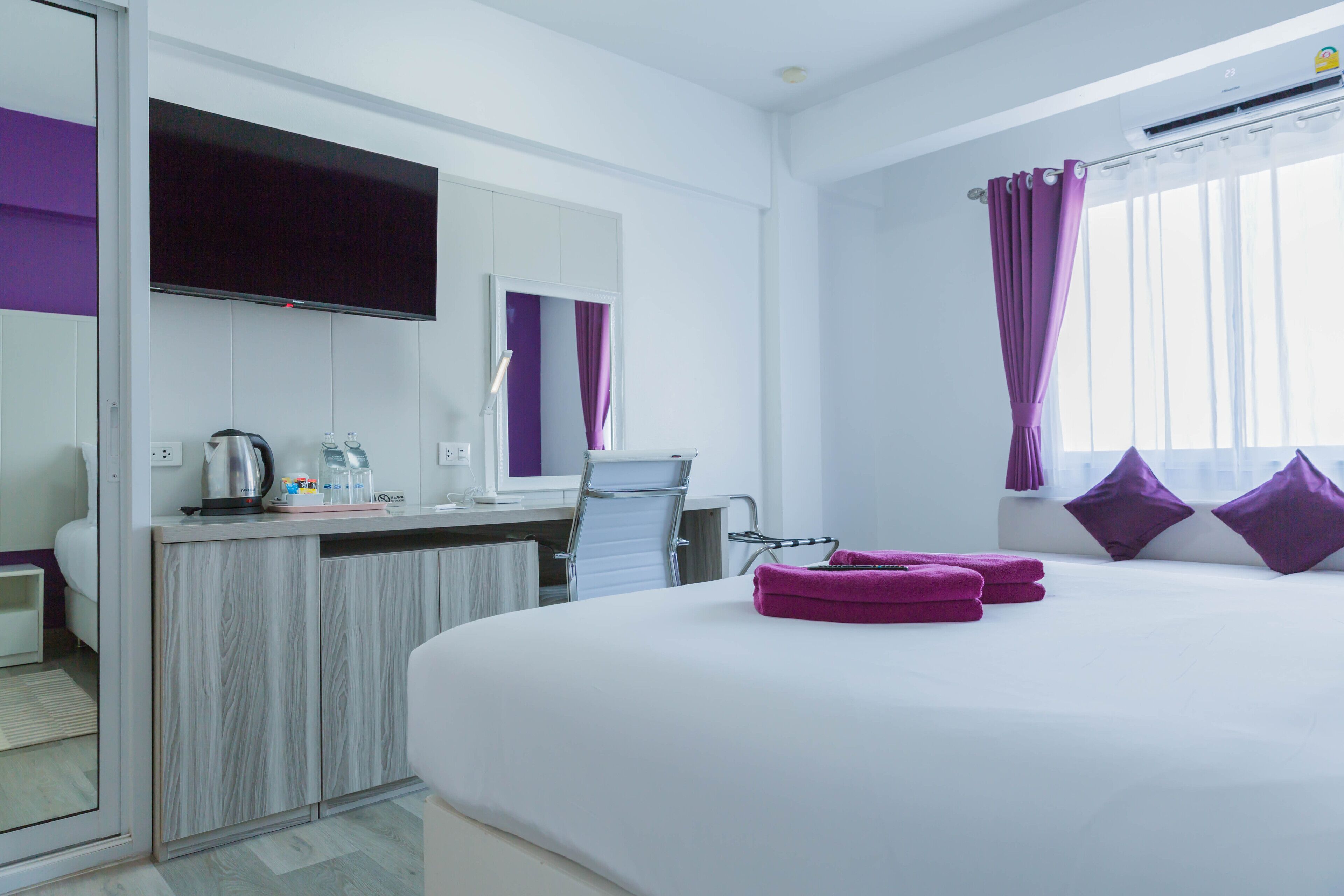 Ein modernes Hotelzimmer mit einem großen Bett, einem Schreibtisch mit Stuhl, einem Flachbildfernseher, einem Fenster mit violetten Vorhängen und einem durchgegangenen Tür sichtbar erkennbaren Badezimmer.