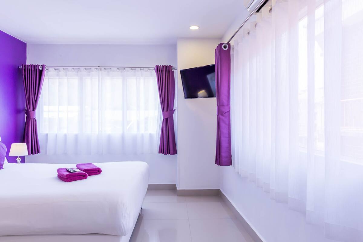 Una habitación de hotel moderna con una cama grande, un televisor de pantalla plana y cortinas moradas.