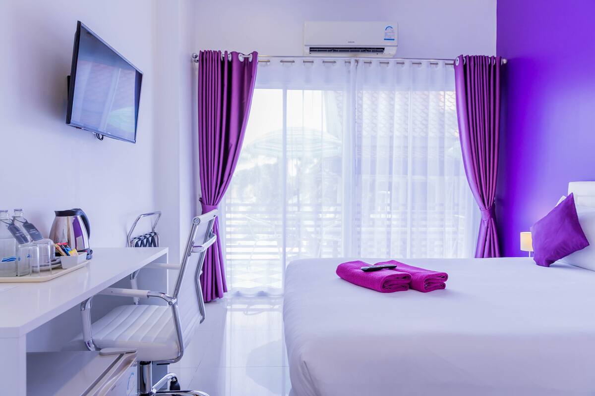 Una habitación de hotel con cama, televisión, escritorio con hervidor y cortinas moradas.