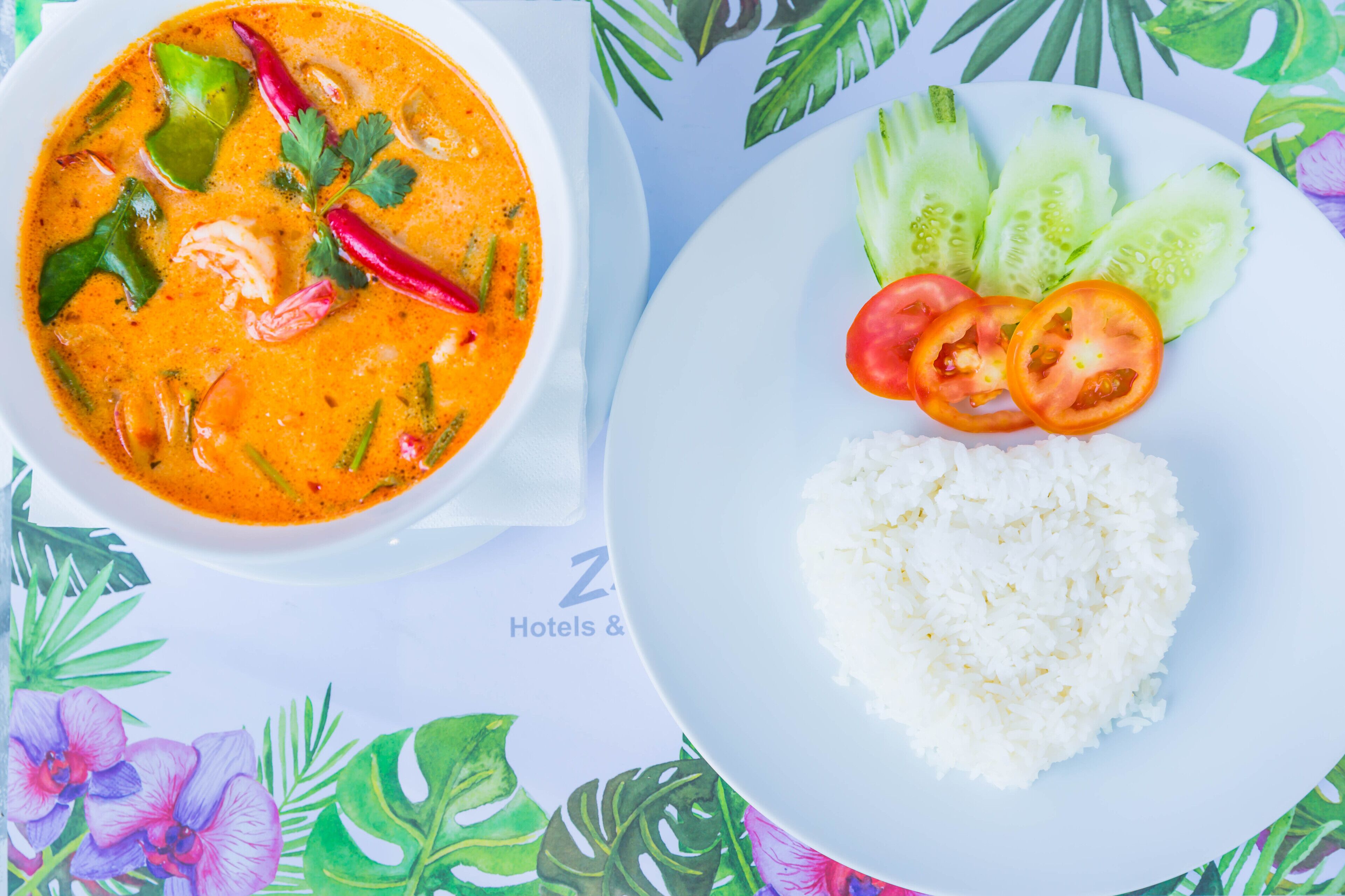 A bowl of soup with vegetables and a plate of rice and cucumber slices.