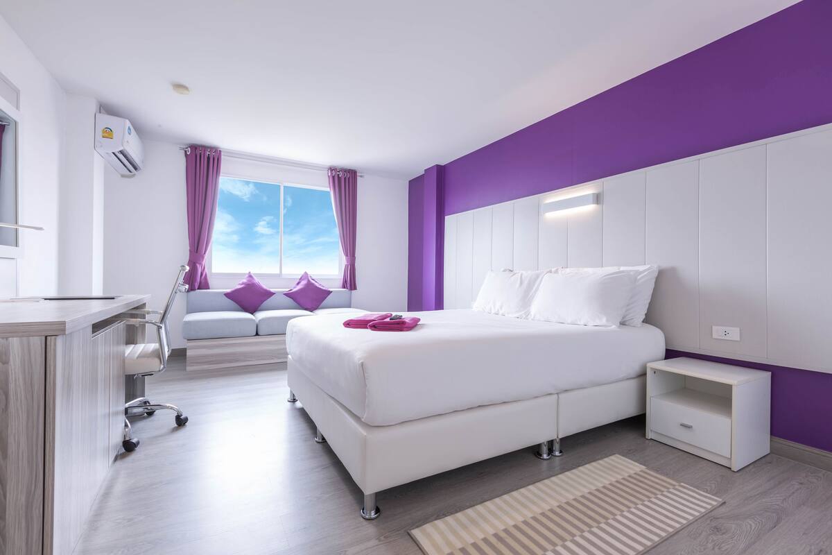 Un dormitorio moderno con una cama blanca, detalles morados, un escritorio y una ventana con cortinas.