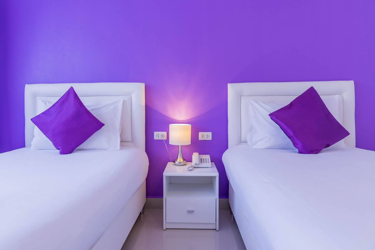Dos camas con cabeceros blancos y almohadas moradas, una mesita de noche con lámpara y teléfono, y una pared morada.