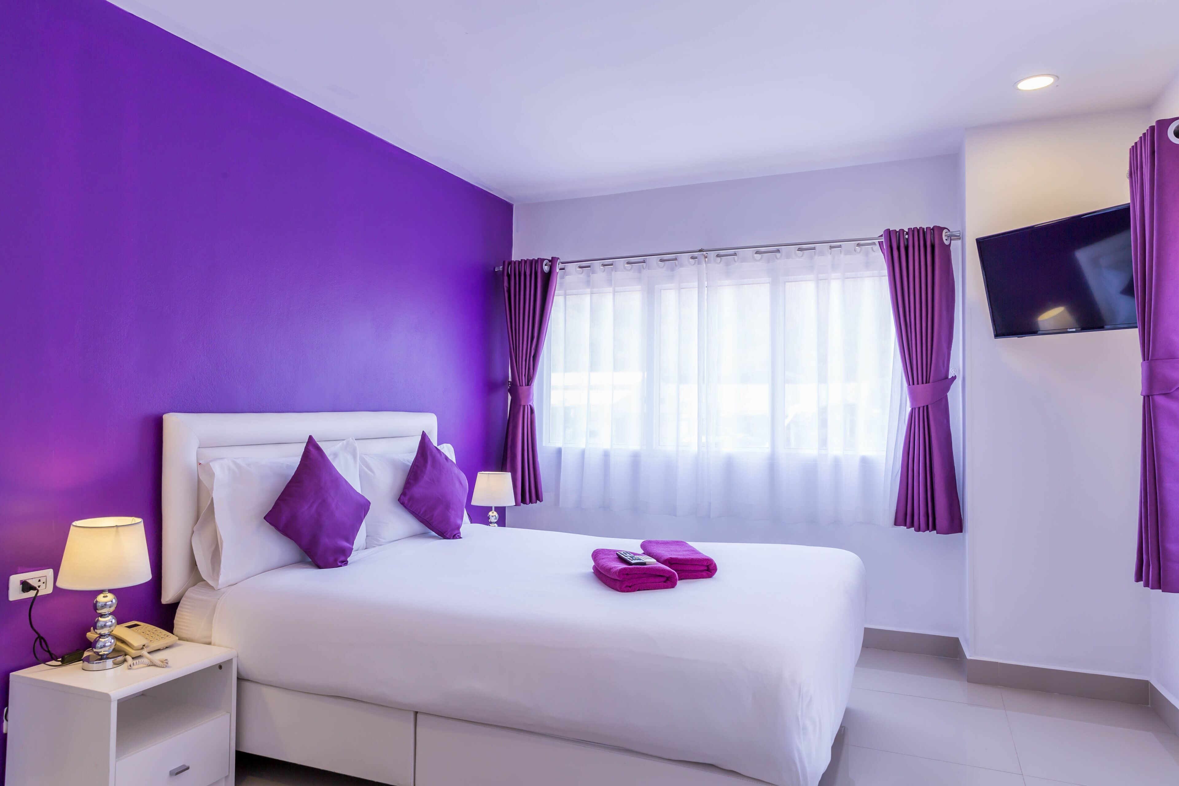 Een hotelkamer met een wit bed, paarse kussens en gordijnen, een flatscreen televisie aan de muur en een raam met dunne gordijnen.